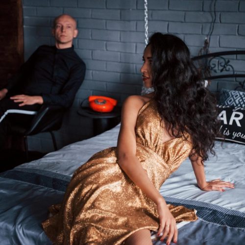 Elegant woman in golden dress sitting on bed with man in background indoors